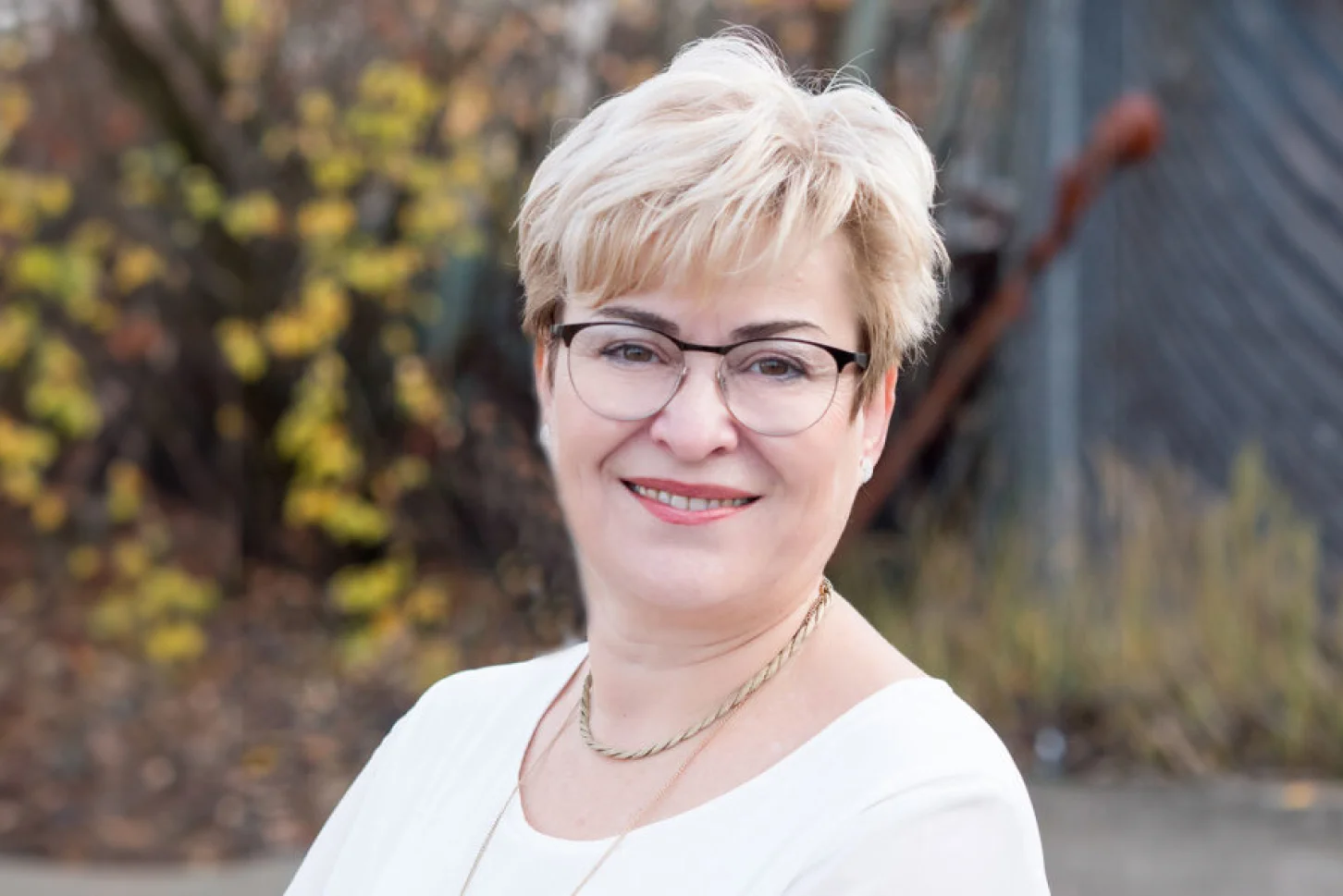 Porträtfoto einer lächelnden Frau mit kurzen blonden Haaren und Brille in weißer Bluse