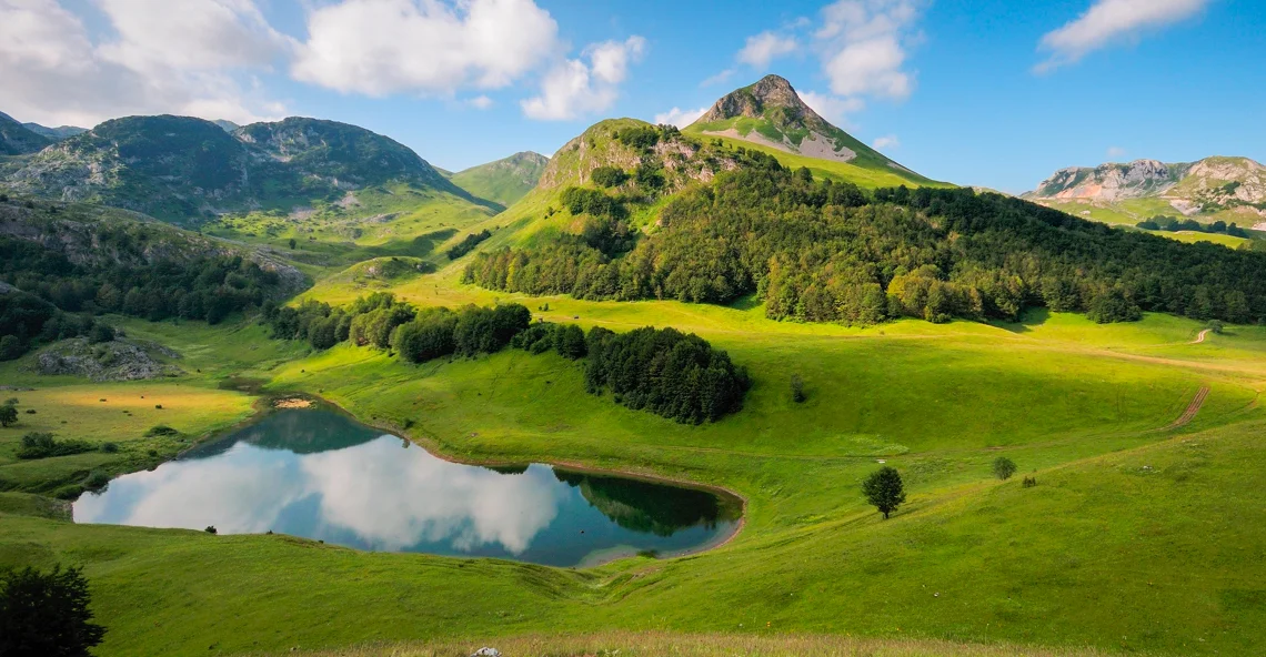 Bergsee umgeben von grünen Wiesen und bewaldeten Hügeln vor blauem Himmel