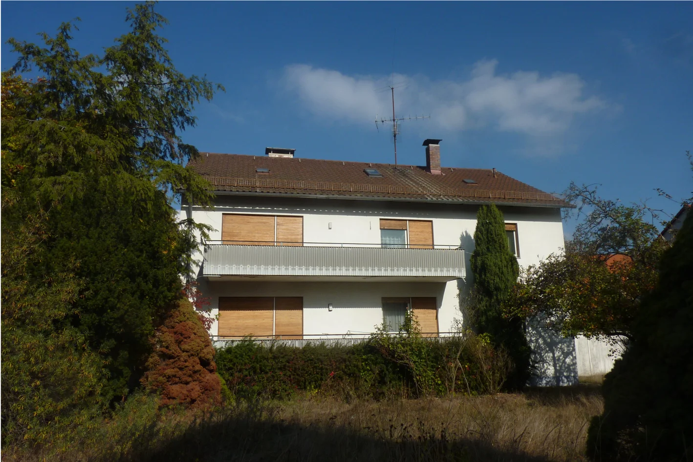 Weißes Haus mit Holz-Fensterläden und Balkon vor blauem Himmel, umgeben von Nadelbäumen