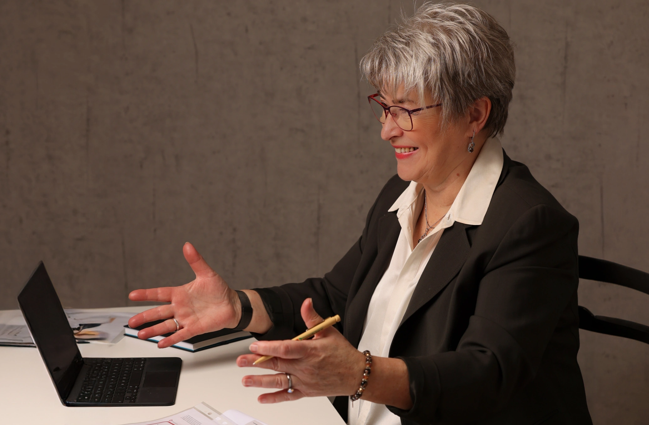 Lächelnde Person am Schreibtisch mit Laptop und Stift, gestikulierend im Gespräch