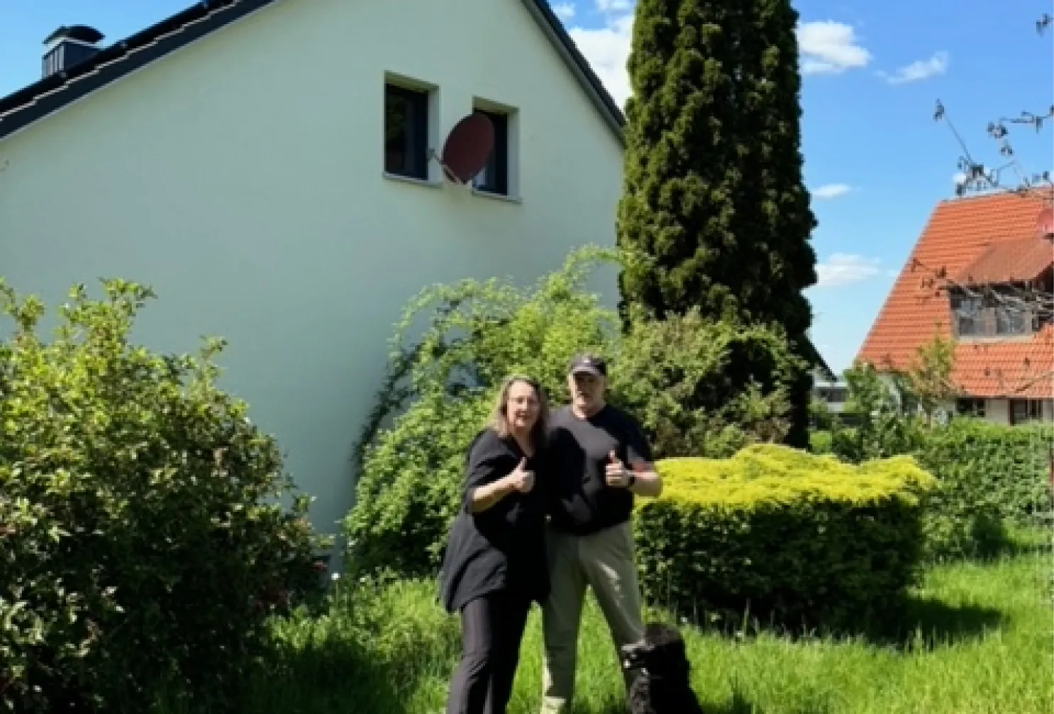 Zwei Personen mit Hund vor einem hellen Wohnhaus in grüner Gartenumgebung