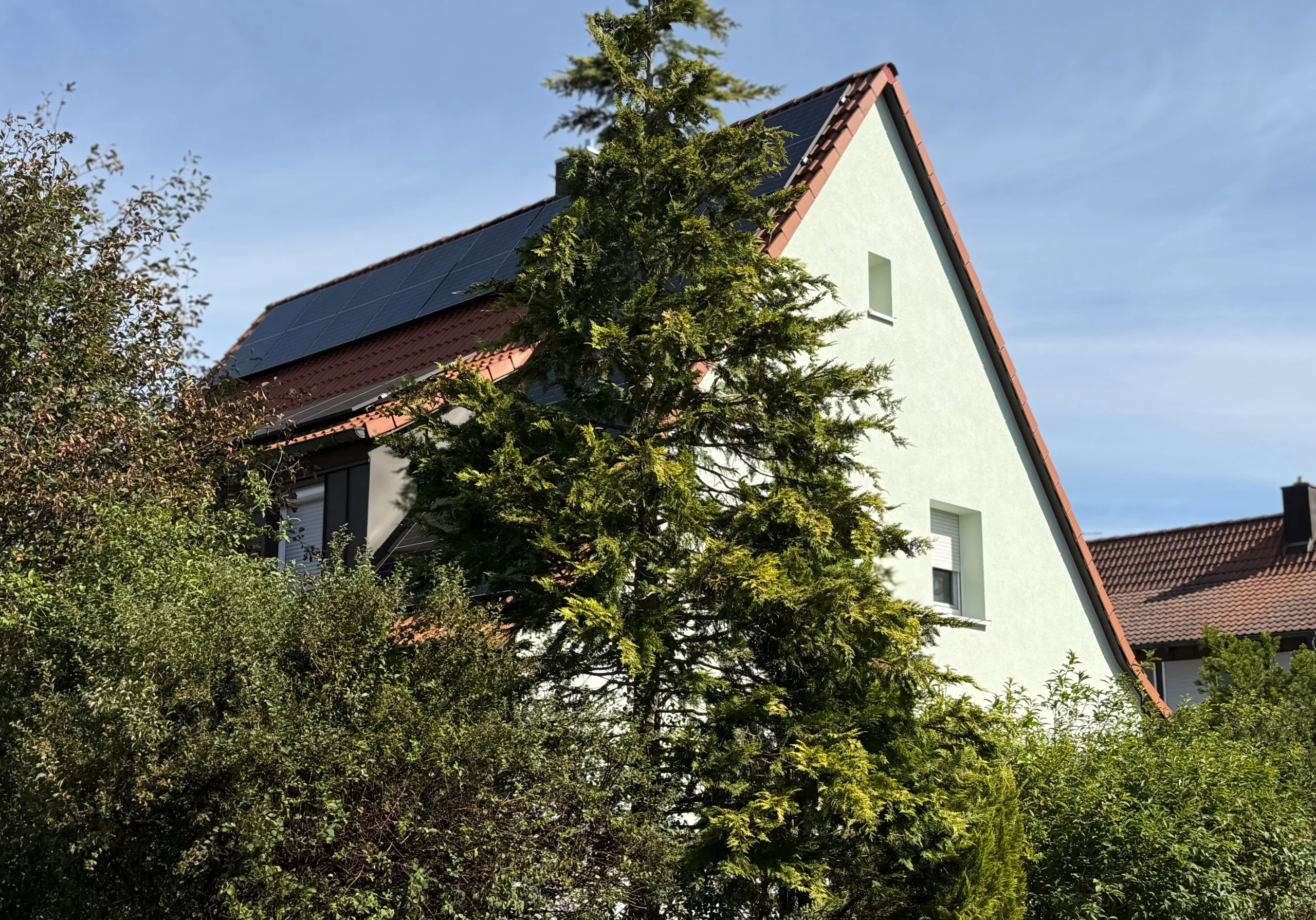Weißes Einfamilienhaus mit Solaranlage auf dem Dach hinter hohen Nadelbäumen