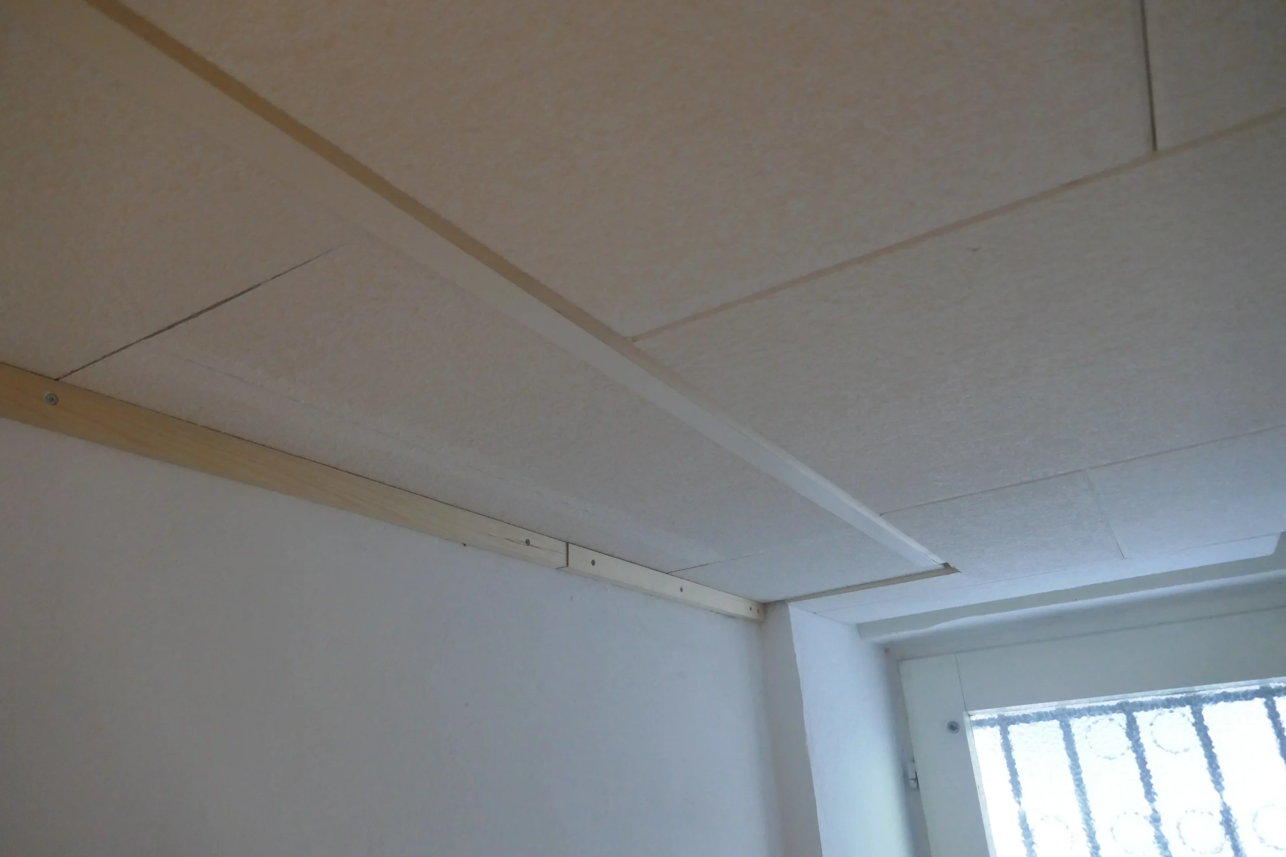 Deckenecke mit weißen Deckenplatten und montierter Holzlatte über einem vergitterten Fenster