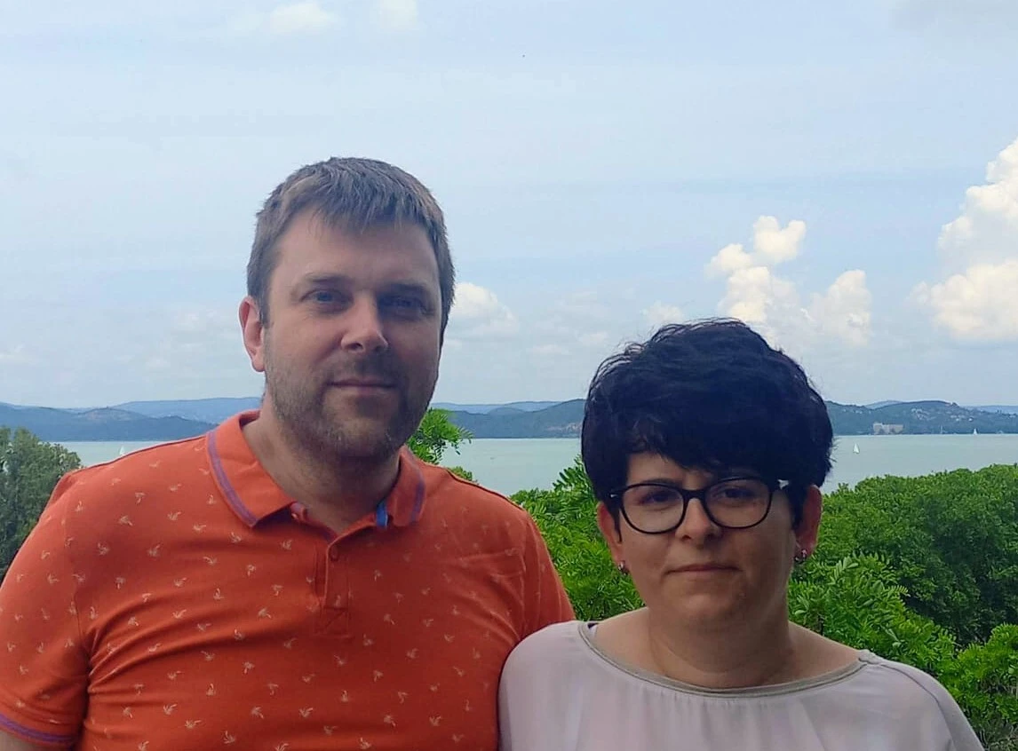 Porträtfoto zweier Personen vor einem See mit Hügeln und Wolken im Hintergrund